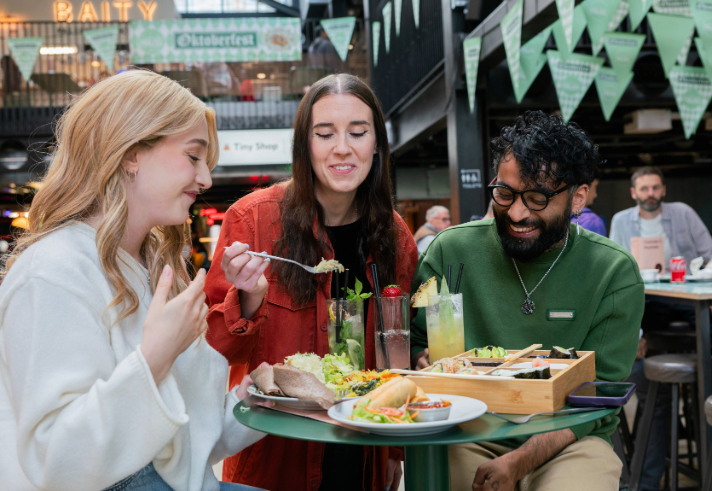 Three people seated at a round green table in an indoor food hall, sharing a meal. The table holds plates of tacos, salad, bread, and a wooden tray with assorted dishes. There are also tall glasses of colourful drinks garnished with fruit and herbs. In the background, green triangular banners hang overhead, and signs for “Oktoberfest” and “Baity” are visible, creating a lively, casual dining atmosphere.