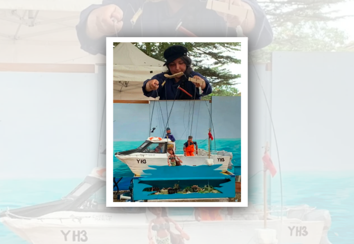 A puppeteer stands behind a small outdoor puppet theatre, manipulating strings attached to a model fishing boat and miniature figures. The scene features a white boat marked “YH3” floating on painted blue water, with puppet characters on board and alongside the vessel. A canopy tent and trees form the background, creating the setting for a live marionette performance.