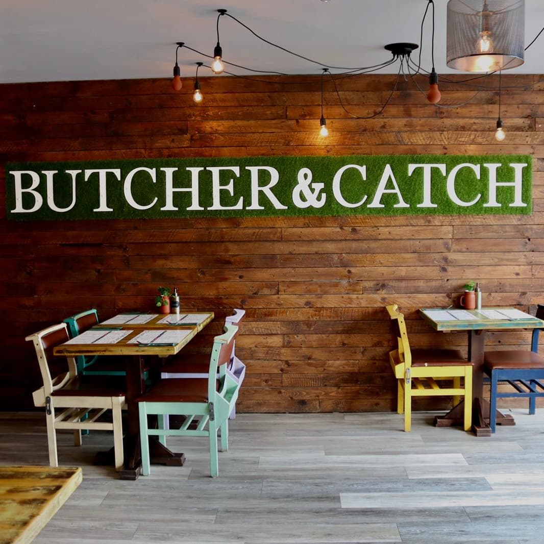 Restaurant tables next to a naked brick wall with a green sign that reads 'Butcher & Catch'.