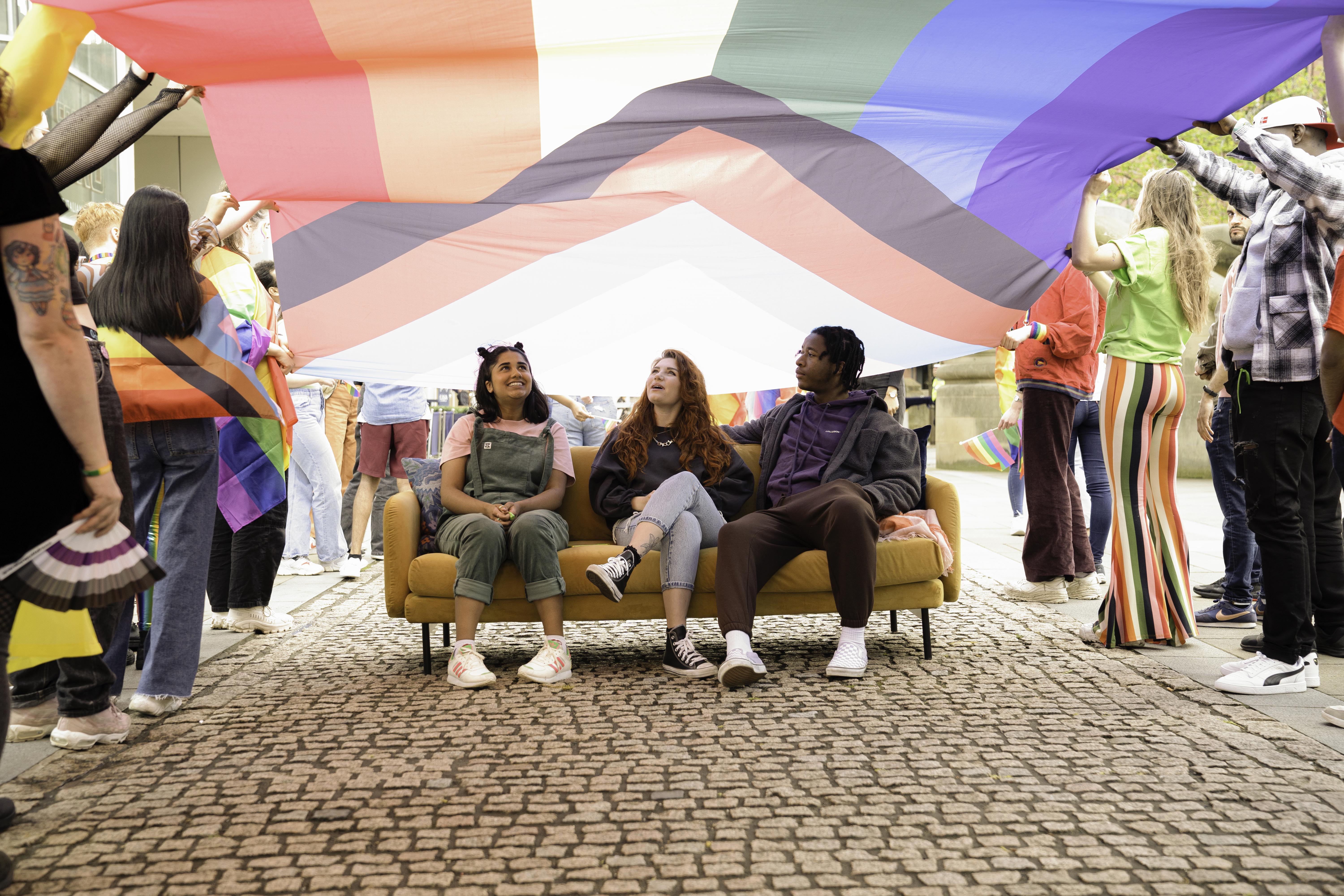 Three people sitting on a yellow sofa under a large Progress Pride flag being held overhead by a group of people. The flag displays rainbow colors along with chevron stripes in black, brown, white, pink, blue, and light blue. The scene takes place outdoors on a cobblestone walkway, with individuals standing around holding sections of the flag.