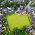 Aerial view of a rectangular green lawn, possibly a bowling green, surrounded by residential houses and narrow streets. The area includes trees, gardens, and parked cars, with a mix of brick buildings and greenery visible throughout the neighbourhood.
