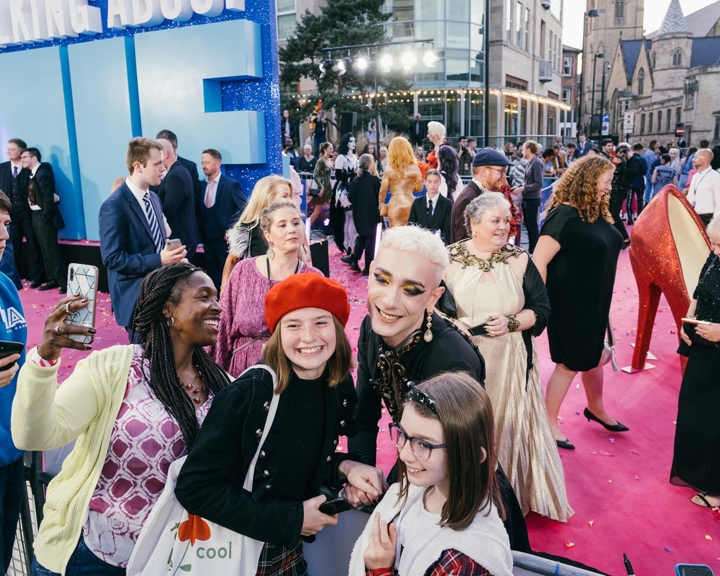 Three people smile and take a selfie together at a red carpet event. Behind them, other attendees in both formal and casual attire mingle. Large blue letters spelling 'ME' are visible, likely part of a sign reading 'EVERYBODY'S TALKING ABOUT JAMIE.' The event takes place outdoors with buildings and lights in the background.