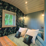 Cosy, stylish bedroom with blue panelled walls and a curved white ceiling. A double bed is dressed in soft blue bedding, large white pillows and patterned cushions matching the dark botanical wallpaper, which features trailing green leaves and small birds. Warm wall-mounted lights create a welcoming atmosphere, while a window frames views of lush green foliage outside.