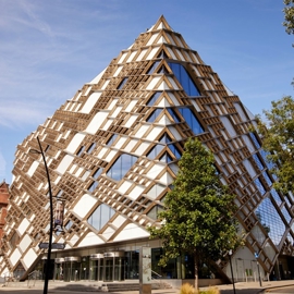 Modern pyramid-shaped building with a striking geometric design featuring glass panels and wooden lattice framework, located on a street corner with trees and older brick buildings nearby under a clear blue sky.