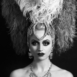 A black‑and‑white portrait of a person wearing an elaborate showgirl-style costume, featuring a large feathered headdress with ornate beading and a tall plume rising from the centre. They also wear a decorative beaded necklace. The background is dark, making the dramatic headdress stand out.