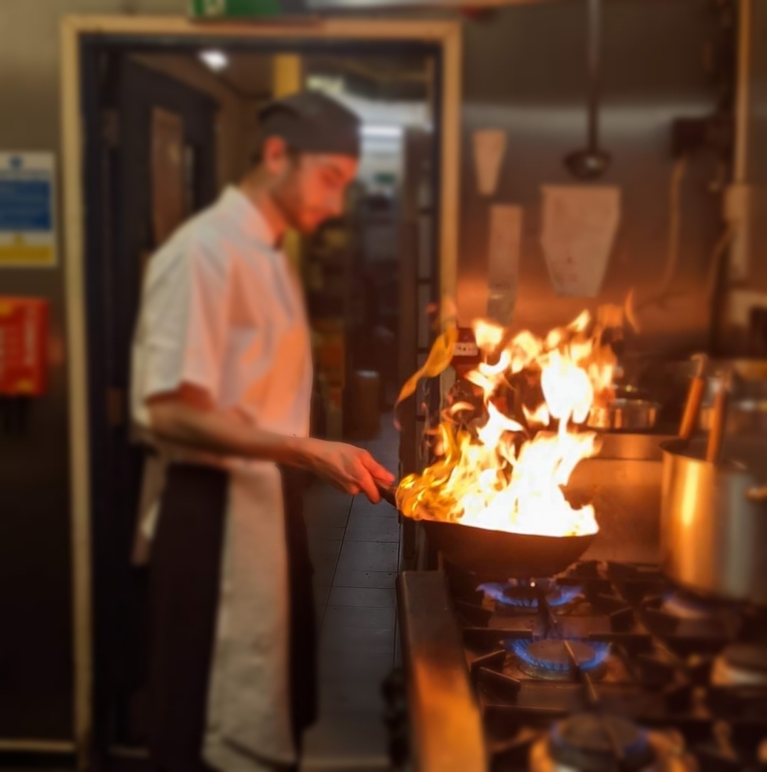 A man cooking with a wok in a kitchen, there are flames leaping from the wok.