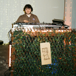 DJ performing behind a mixing desk at an indoor event venue. The booth is decorated with leafy green garlands and warm strip lighting, while fairy lights hang across the backdrop. Audio equipment and turntables sit on the table, and colourful artwork is visible to one side. The industrial-style space features concrete floors and ambient lighting, creating a relaxed atmosphere for live music and entertainment.