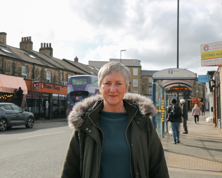Person in a dark green jacket with a fur-lined hood standing on a city sidewalk. Behind them, a purple double-decker bus approaches a bus stop where people are waiting. Urban buildings, parked cars, and pedestrians line the street under a partly cloudy sky.