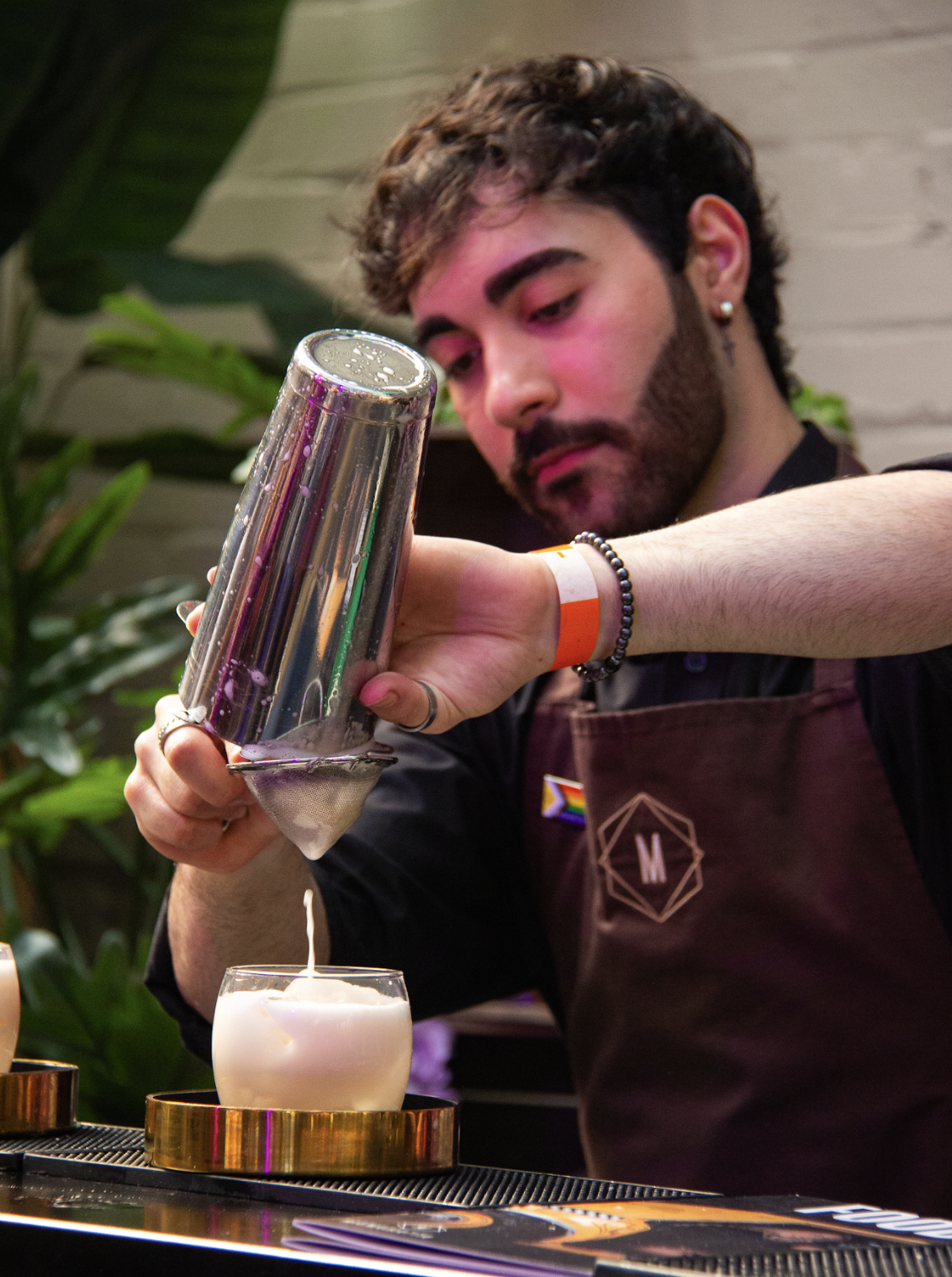 A bartender pours a creamy white drink from a metal shaker into a round glass set on a gold coaster. They wear a dark apron with a geometric logo and a small pride flag badge. Green plants and soft lighting create a vibrant, welcoming bar setting.