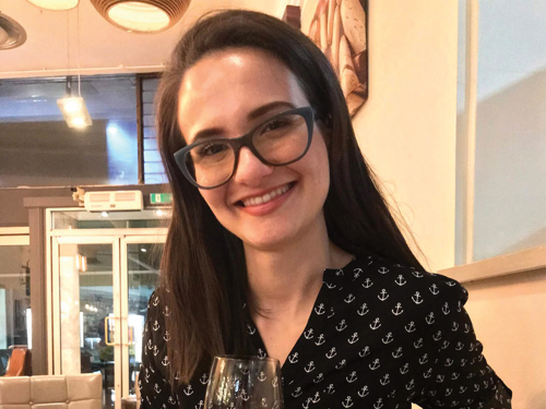 A person seated indoors at a restaurant table, holding a wine glass. The individual is wearing a black shirt patterned with small white anchor designs. The background shows modern décor with wooden panels, ceiling lights, and a glass door leading outside.