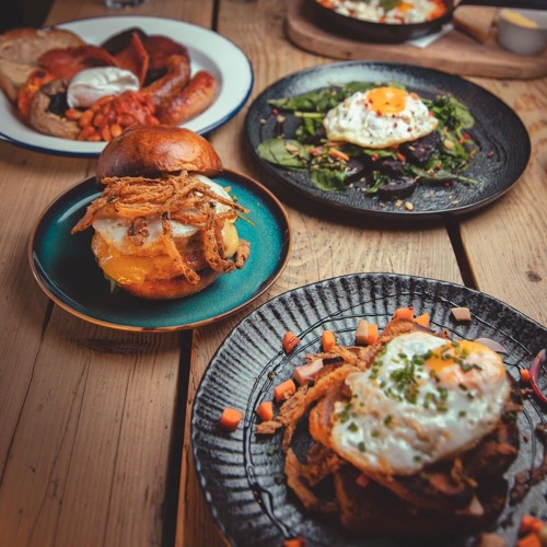 A wooden table set with four plates of hearty breakfast dishes. In the foreground, a black plate holds a toasted sandwich topped with a fried egg, crispy onions, and garnished with herbs and diced vegetables. To the left, a brioche bun sandwich filled with melted cheese, fried egg, and crispy onions sits on a teal plate. At the back, a plate features a traditional English breakfast with sausages, bacon, beans, and a fried egg. Another black plate displays a bed of greens topped with a sunny-side-up egg. The scene is warmly lit, highlighting the rich colours and textures of the food.