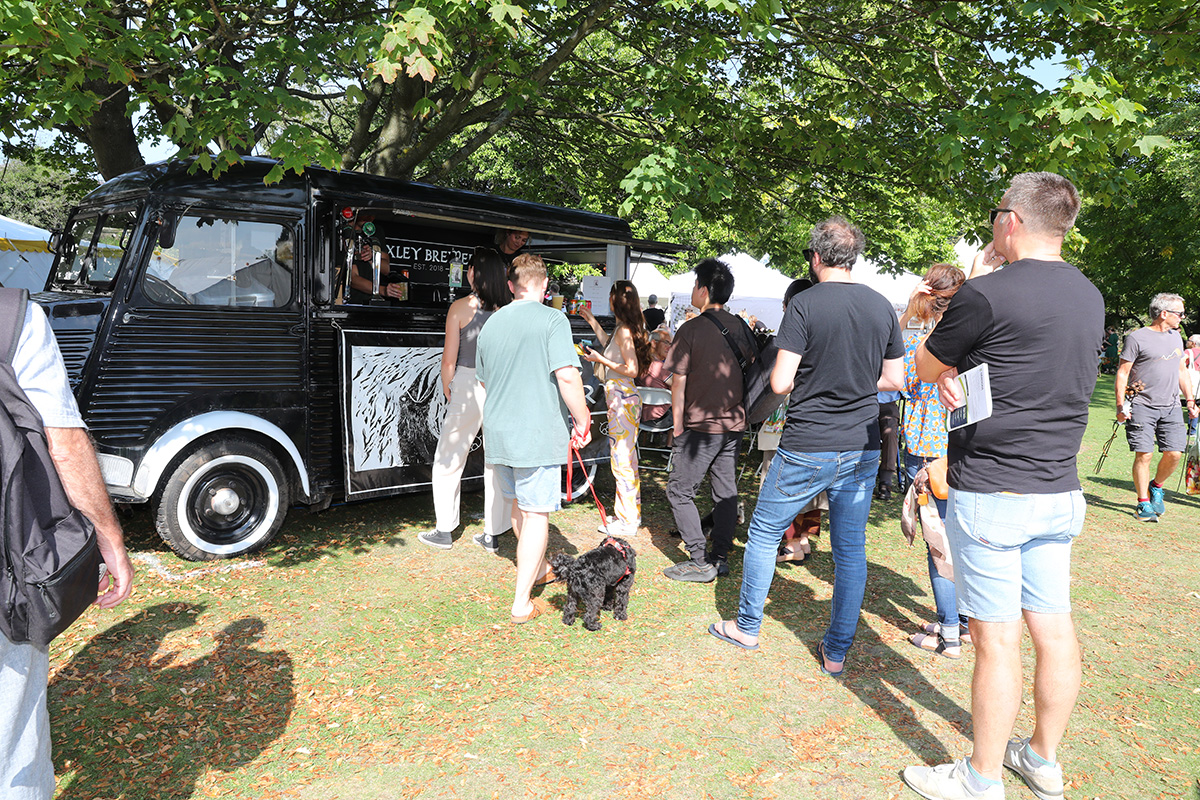 A black vintage-style food and drink van with “Loxley Brewery” signage serves customers at an outdoor event. Several people are queuing on the grass under leafy trees, some holding drinks and others accompanied by dogs. The scene is bright and sunny, with white tents visible in the background, suggesting a festival or garden fair setting.