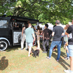 A black vintage-style food and drink van with “Loxley Brewery” signage serves customers at an outdoor event. Several people are queuing on the grass under leafy trees, some holding drinks and others accompanied by dogs. The scene is bright and sunny, with white tents visible in the background, suggesting a festival or garden fair setting.