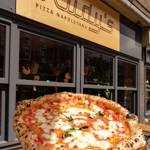 A freshly baked Neapolitan pizza with melted mozzarella, tomato sauce and basil held up in front of a restaurant exterior with a sign reading “Rudy’s Pizza Napoletana.”