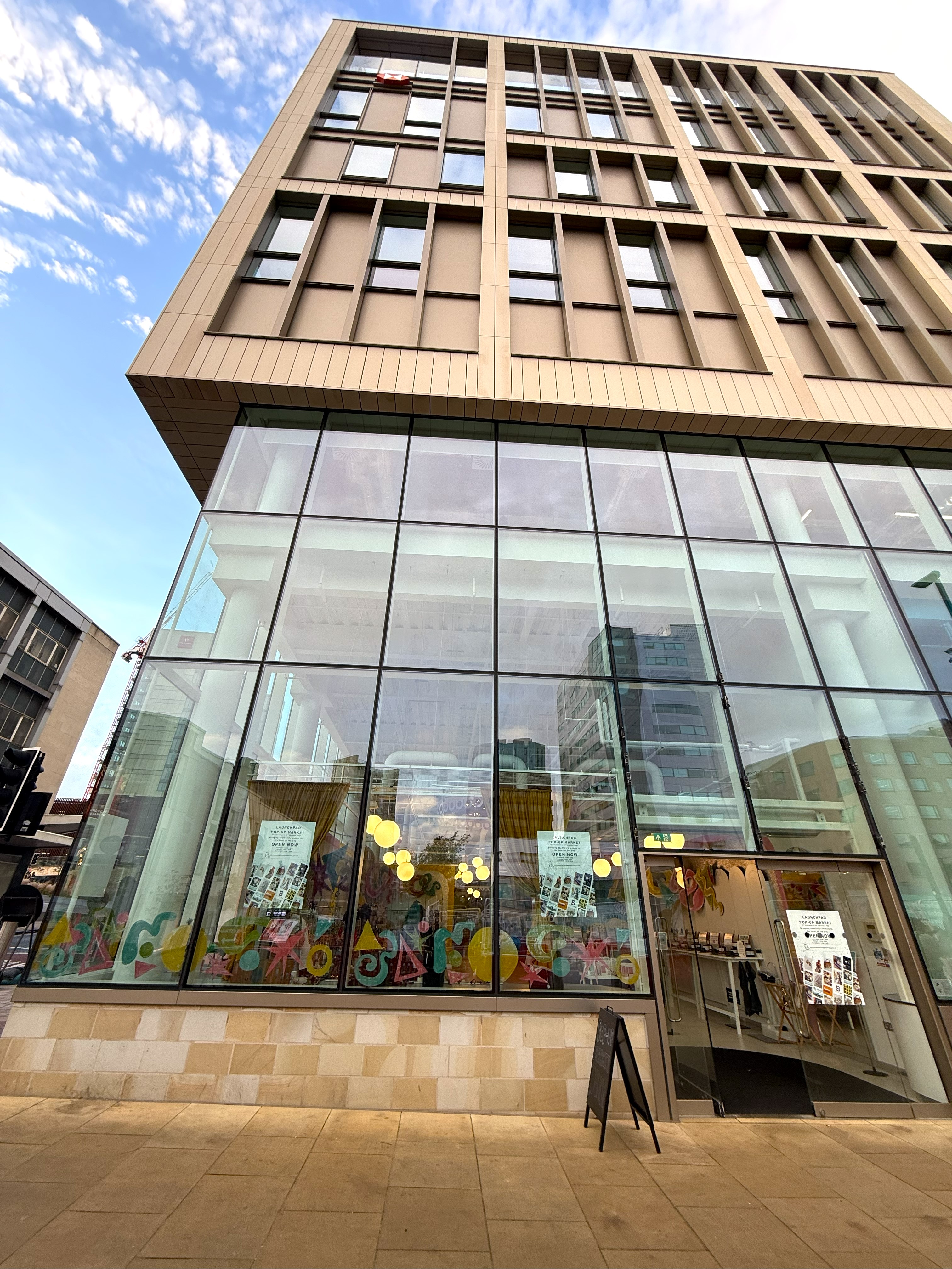 Exterior view of a modern multi-story building with large glass windows showcasing a colorful pop-up market inside. Posters on the windows promote the Launchpad Pop-Up Market, and a black sidewalk sign near the entrance reads 'Pop-Up Market Open Now.