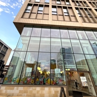 Exterior view of a modern multi-story building with large glass windows showcasing a colorful pop-up market inside. Posters on the windows promote the Launchpad Pop-Up Market, and a black sidewalk sign near the entrance reads 'Pop-Up Market Open Now.
