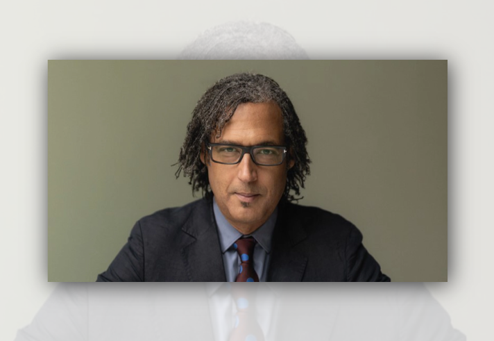 tudio portrait of a person wearing a dark suit, blue shirt and patterned tie, seated against a plain grey background. The person has shoulder-length textured hair and rectangular glasses, looking directly towards the camera. Soft, even lighting highlights the face, creating a professional and formal appearance.