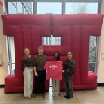 Four people stand in front of a huge inflatable letter 'H'. One of them is holding a pink t-shirt with the St Luke's logo on it.