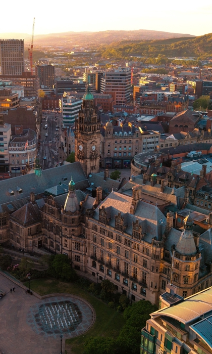 Aerial view of Sheffield city centre at golden hour, featuring the historic Town Hall with its clock tower and surrounding civic buildings, with modern offices, streets, and green hills stretching into the distance.