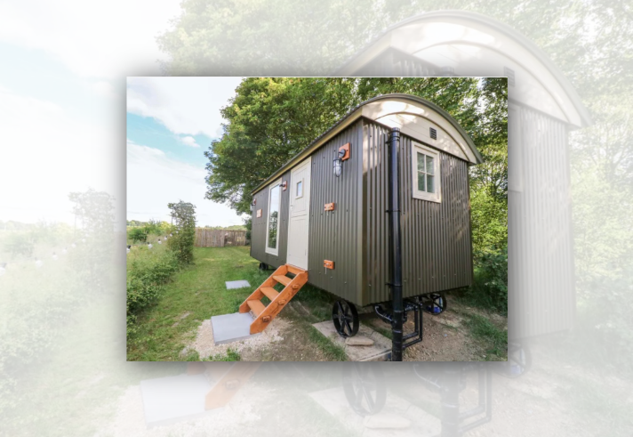 Exterior of a compact shepherd’s hut with dark green corrugated metal cladding and a curved cream roof, set in a grassy countryside location. Wooden steps lead to a white door, with windows on the side and end walls. Surrounded by mature trees and open greenery, the hut sits on traditional wagon-style wheels, creating a peaceful rural retreat.