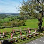 A wide view of The Old Horns Inn beer garden overlooking rolling green countryside, with rows of picnic tables and red parasols set against a backdrop of fields and hills.