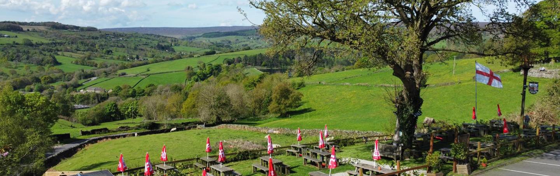 A wide view of The Old Horns Inn beer garden overlooking rolling green countryside, with rows of picnic tables and red parasols set against a backdrop of fields and hills.