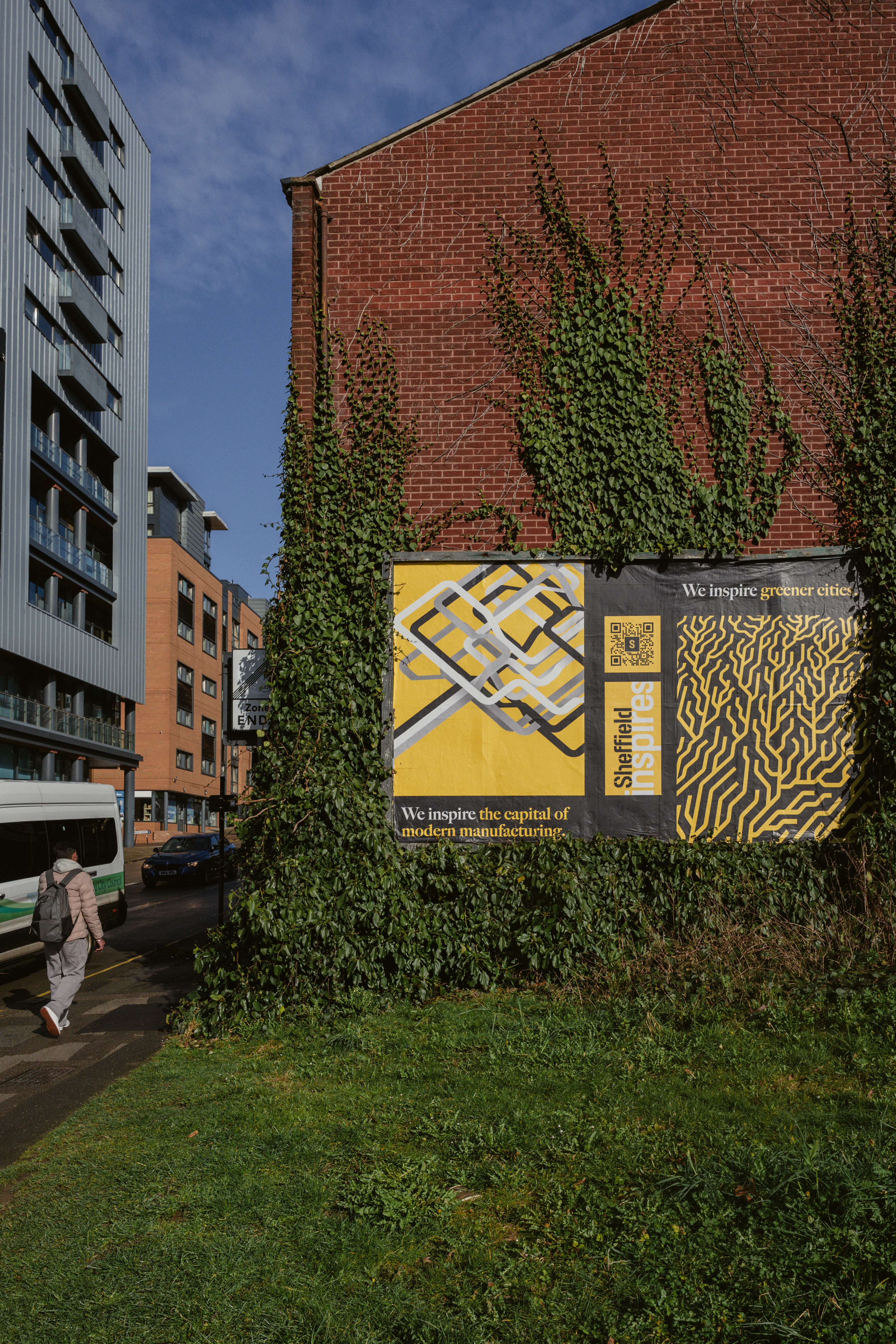 A large brick wall partially covered with green ivy, featuring two posters with bold yellow and black designs. The left poster shows an abstract geometric pattern with the text “We inspire the capital of modern manufacturing.” The right poster has an organic, maze-like pattern with the text “We inspire greater cities.” Both posters include the phrase “Sheffield inspires” and a QR code. In the foreground, there is a grassy area, and to the left, a person walks along the pavement near a white and green bus. 
