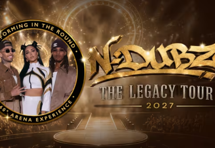 Promotional graphic for “N-Dubz: The Legacy Tour 2027”. A gold and bronze arena-themed background features a circular stage and concert lighting. On the left, a round frame shows three performers in coordinated stage outfits beneath the text “Performing in the Round” and “360° Arena Experience”. Large gold lettering across the centre reads “N-Dubz” with “The Legacy Tour 2027” underneath. The design uses metallic effects and bright spotlight accents.
