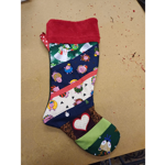 Handmade Christmas stocking made from patchwork fabric panels, photographed on a wooden tabletop. The stocking features a red fleece cuff and colourful festive prints including cartoon pigs in Christmas outfits, a snowman, stars, trees, and a bird. Decorative diagonal strips, a red polka-dot ribbon loop, and an appliqué heart add detail. Small fabric trimmings are scattered on the work surface around the stocking.