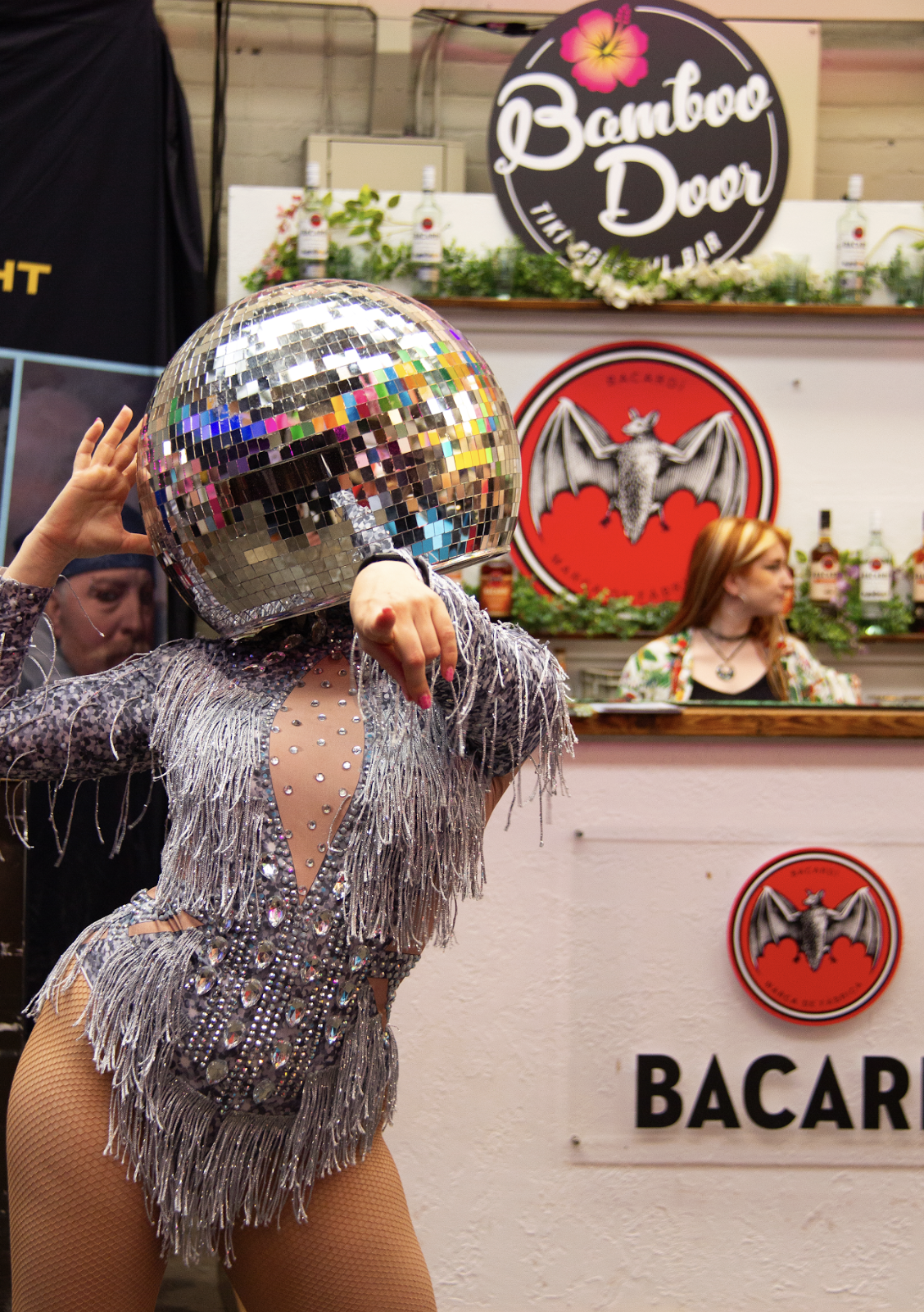 A performer dressed in a sparkling, silver fringed bodysuit poses with one arm raised and the other pointing forward. They wear a large disco‑ball helmet that reflects colourful light. Behind them is a bar area decorated with greenery, branded signs and a circular “Bamboo Door” logo, creating a lively, festive atmosphere.