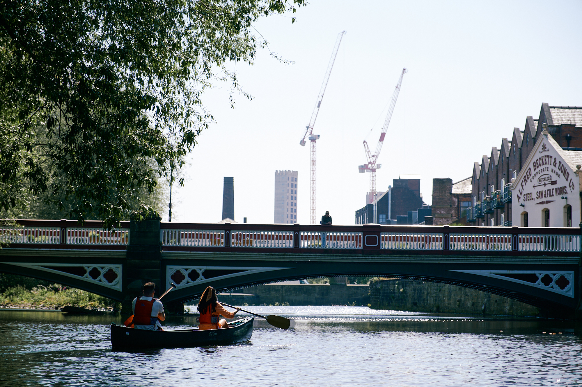 Couple kayaking in Kelham Island on the River Don towards Ball Street Bridge, with city view in the background 