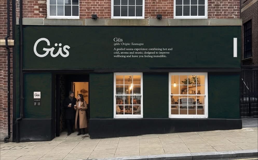 Street view of a dark green storefront with the name ‘Güs’ in large white letters on the upper left. To the right, text describes Güs as ‘A guided sauna experience combining hot and cold, aroma and music; designed to improve wellbeing and leave you feeling incredible.’ Two large windows reveal a cozy interior with warm lighting and shelves. A doorway on the left shows two people exiting, one holding a drink. The building has a brick upper facade and sits on a paved sidewalk.