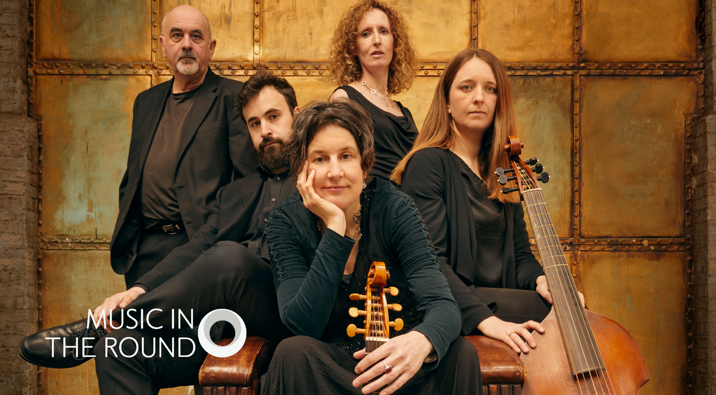 Five musicians dressed in black sit and stand against a rustic golden metal backdrop. They hold period instruments, including a viola da gamba and a lute, suggesting a classical or early music ensemble. The logo “Music in the Round” appears in the bottom left corner in white text with a circular design element.