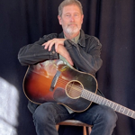 Person seated on a chair in front of a dark curtain backdrop, resting folded arms on an acoustic guitar positioned across their lap. Sunlight from a nearby window casts a bright rectangular pattern on the floor and lower curtain, creating a dramatic contrast with the dark surroundings. The guitar has a sunburst finish and is the central focus of the portrait.