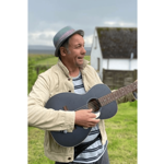 Promotional photograph of musician Martin Stephenson standing outdoors in a grassy rural setting, holding and playing a dark acoustic guitar. Martin wears a light-coloured jacket, striped shirt and grey hat. A white building with a slate roof sits in the background beneath an overcast sky, with green fields stretching into the distance. The image has a soft-focus background that draws attention to the performer and guitar.