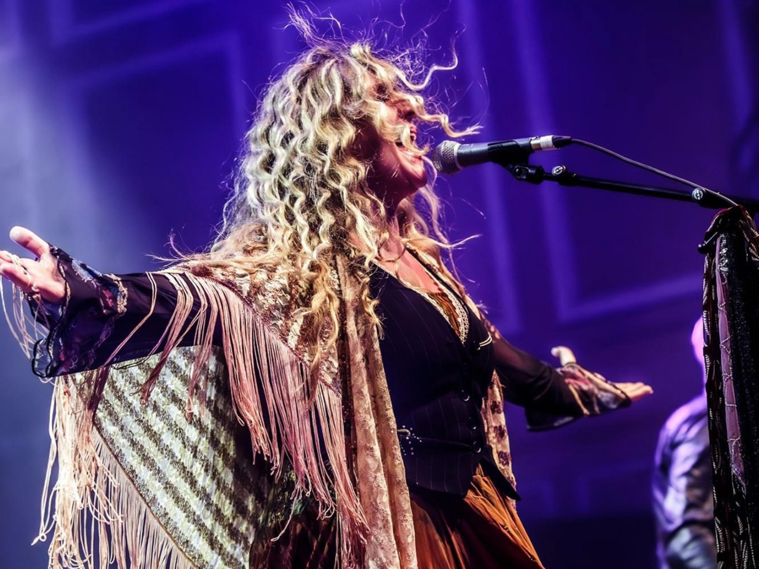 A performer sings passionately into a microphone on stage, arms outstretched, wearing a flowing fringed shawl and layered outfit. Long curly hair catches the stage lights, creating a dramatic effect against a purple-lit background.