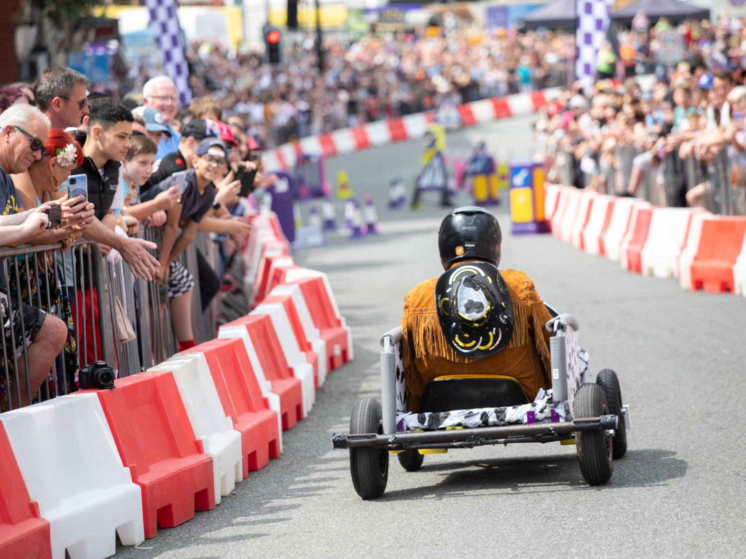 A soapbox cart with a simple frame and four wheels races down a city street lined with red-and-white barriers. The driver wears a black helmet and a fringed brown jacket with a decorative design on the back. Spectators crowd behind metal railings on both sides, watching the event. Purple-and-white race markers and colourful props are visible further down the track.