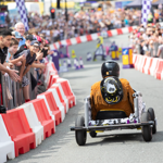 A soapbox cart with a simple frame and four wheels races down a city street lined with red-and-white barriers. The driver wears a black helmet and a fringed brown jacket with a decorative design on the back. Spectators crowd behind metal railings on both sides, watching the event. Purple-and-white race markers and colourful props are visible further down the track.