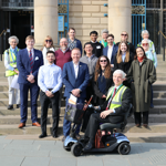 Group photo of around 18 people standing on the steps outside a large civic building with stone columns. The group includes individuals of different ages, some wearing business attire, name badges or high-visibility vests. In the foreground, a man in a mobility scooter smiles at the camera. The group is gathered for a formal visit, event or community engagement activity and poses together in bright daylight.
