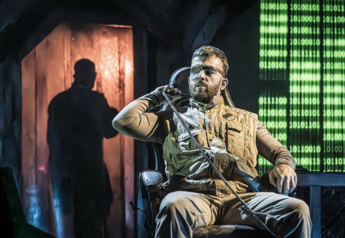  A dramatic stage scene features a person wearing a tactical-style beige outfit and eye patch, seated in a chair under theatrical lighting. A rope is draped across one shoulder. Behind, a shadowy figure stands in a doorway illuminated by orange light. To the right, a glowing digital-style backdrop displays vertical streams of green binary code. The set combines industrial and cyber-themed elements, creating a tense, futuristic atmosphere.