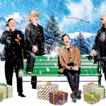 Four people pose in a festive winter-themed scene with falling snow effects. One sits on a green park bench while the others stand nearby. Wrapped gift boxes are arranged around the bench. A vintage payphone stands on the left and a red fire hydrant on the right. The backdrop shows snow-covered mountains, pine trees and a sleigh silhouette in the sky, creating a playful holiday atmosphere.