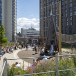 An urban playground surrounded by tall modern buildings, with people sitting on curved benches and children playing on wooden climbing structures and slides. The area includes a rope bridge, landscaped flower beds with colorful blooms, and paved walkways. The background shows city architecture under a bright blue sky with scattered clouds.