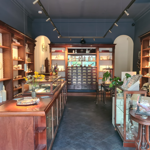 Interior of The Alchemist Gallery, a narrow exhibition and retail space with dark blue walls and polished wooden cabinetry. Glass display cases run through the centre, holding jewellery, ceramics and small artworks. Shelving along both walls displays decorative objects and sculptures. At the back, a vintage apothecary-style cabinet with many labelled drawers forms a focal point. Fresh flowers, plants and spot lighting create a warm, curated atmosphere.