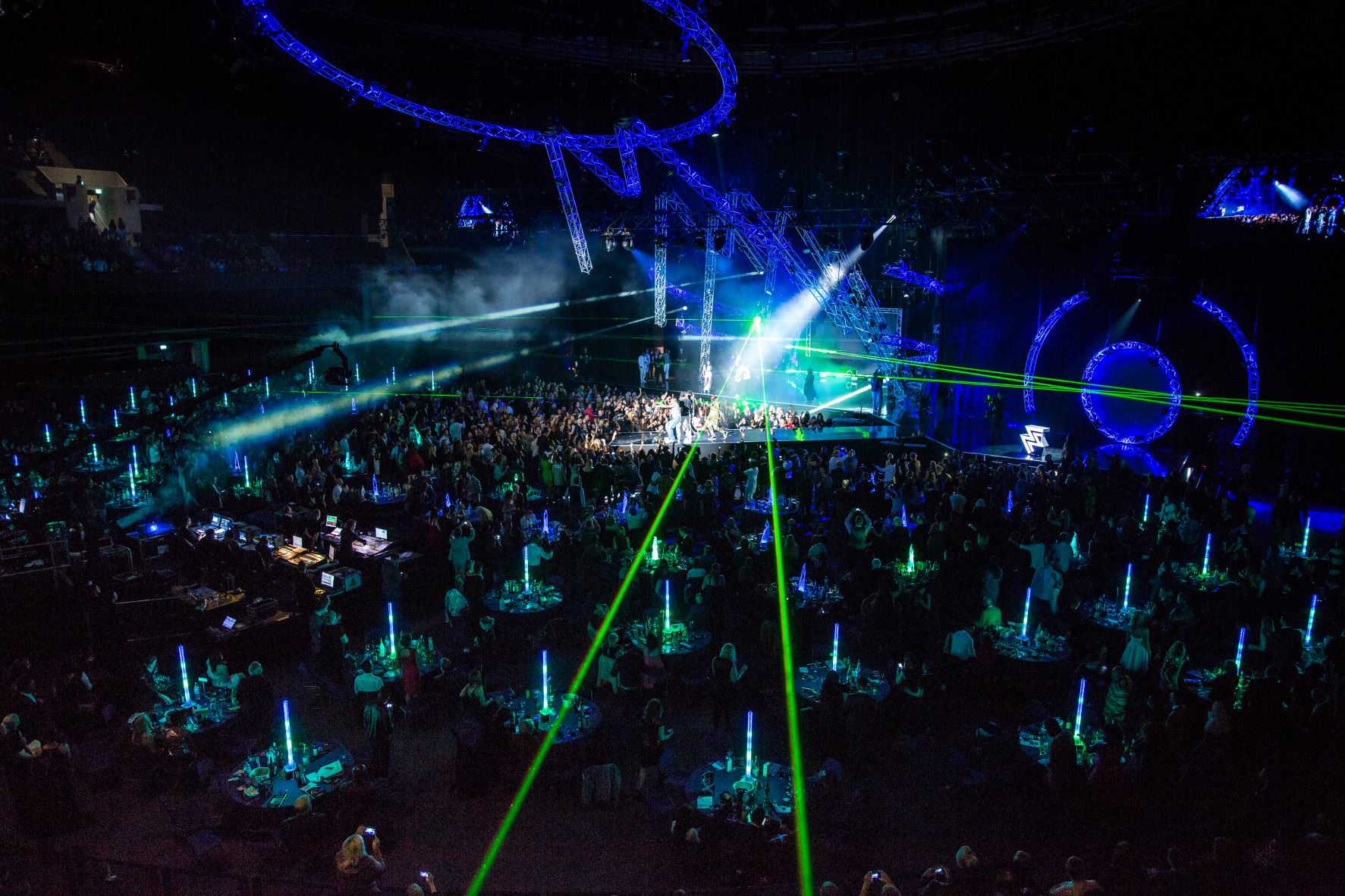 Large arena filled with round tables and seated guests, illuminated by green laser beams and blue accent lighting. A central stage with performers is surrounded by bright spotlights and futuristic light structures, creating a vibrant concert or awards show atmosphere.