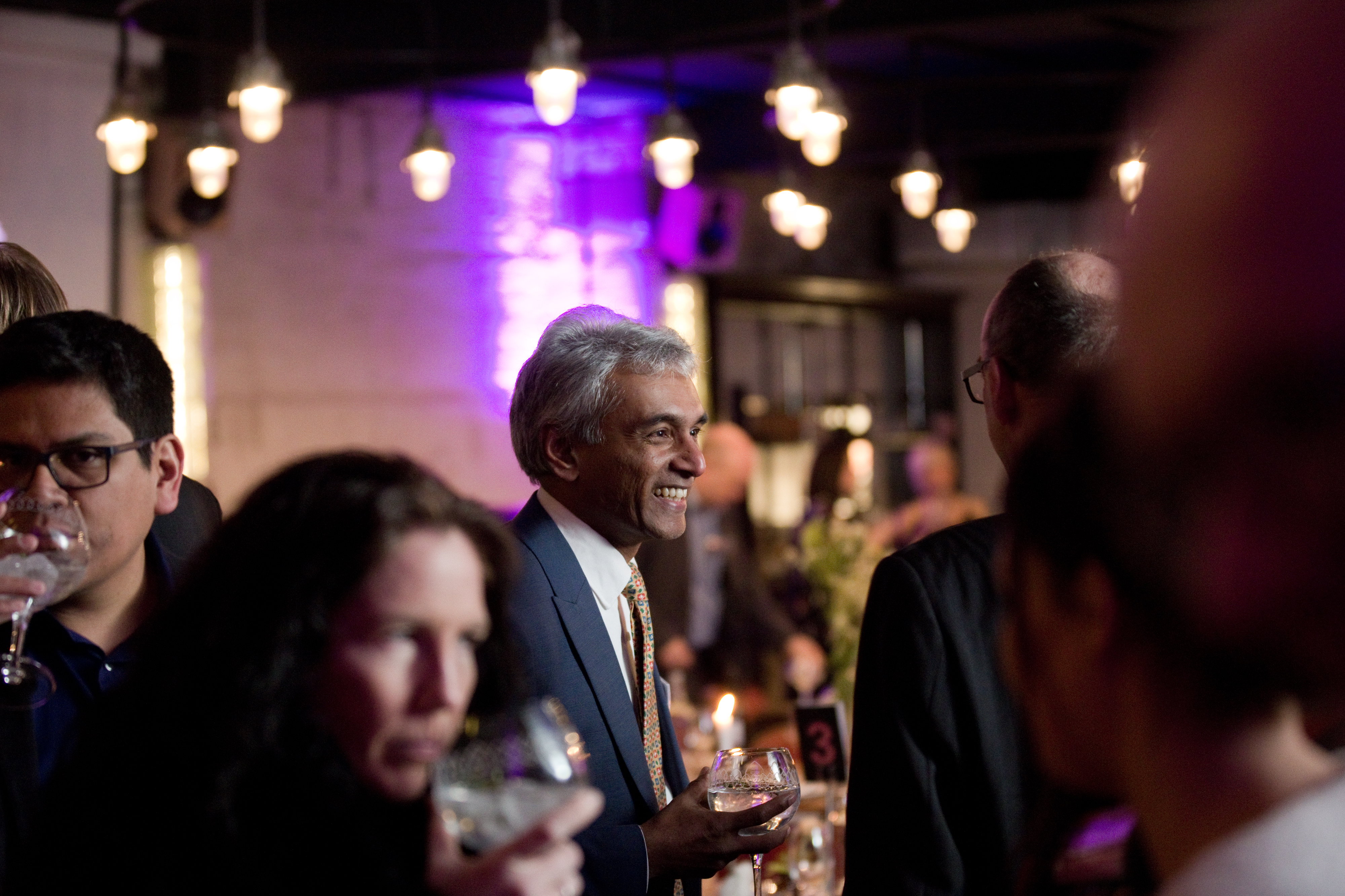 Indoor networking event with people standing and holding drinks in a warmly lit venue. The background features hanging pendant lights, purple accent lighting on the wall, and tables with candles and floral arrangements. A table marker with the number 3 is visible among the decor.
