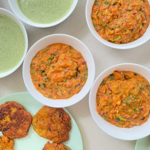 Overhead view of several bowls of food arranged on a light-coloured surface. White bowls contain an orange vegetable-based dip or salad flecked with green herbs, alongside bowls of smooth pale green soup or sauce. Plates at the bottom of the image hold golden-brown fried fritters or patties. The contrasting orange and green colours create a vibrant presentation of freshly prepared dishes.