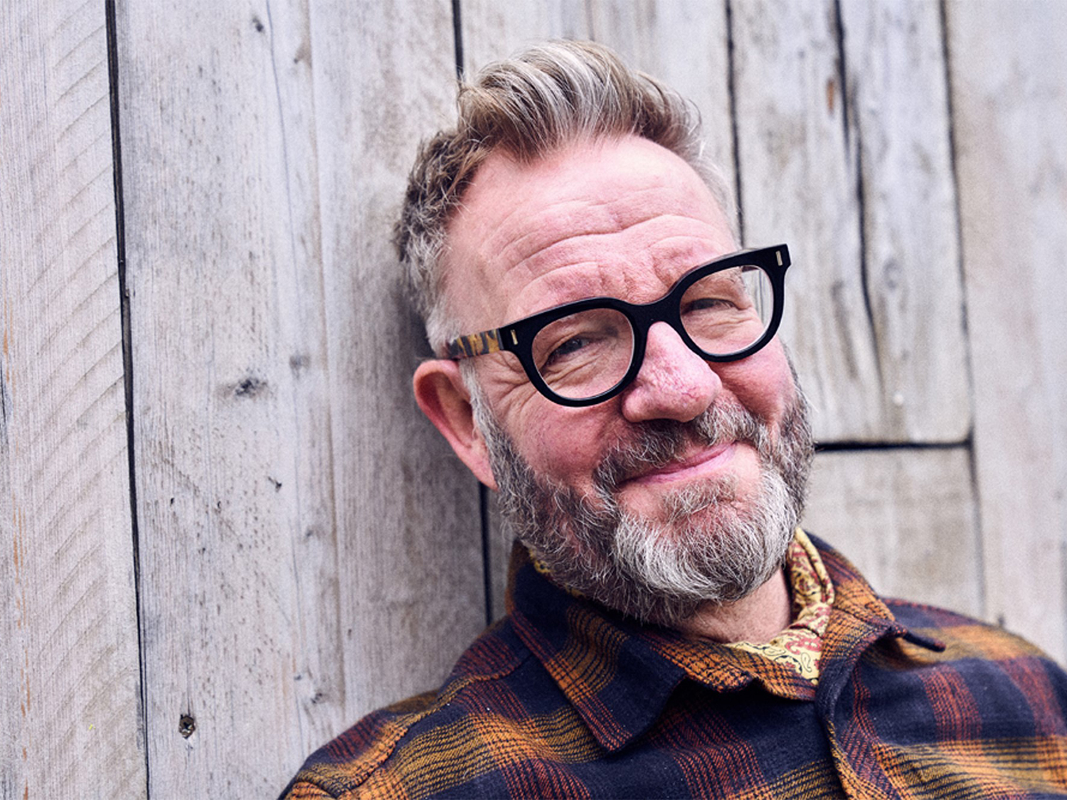 The artist Pete Mckee leaning against a wooden fence, smiling at the camera.