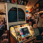 Rutland Arms jukebox lit up, with a chalk board above that's separated into forbidden and recommended. Groups of people are sat to the left of the jukebox, with the bar to the right. 
