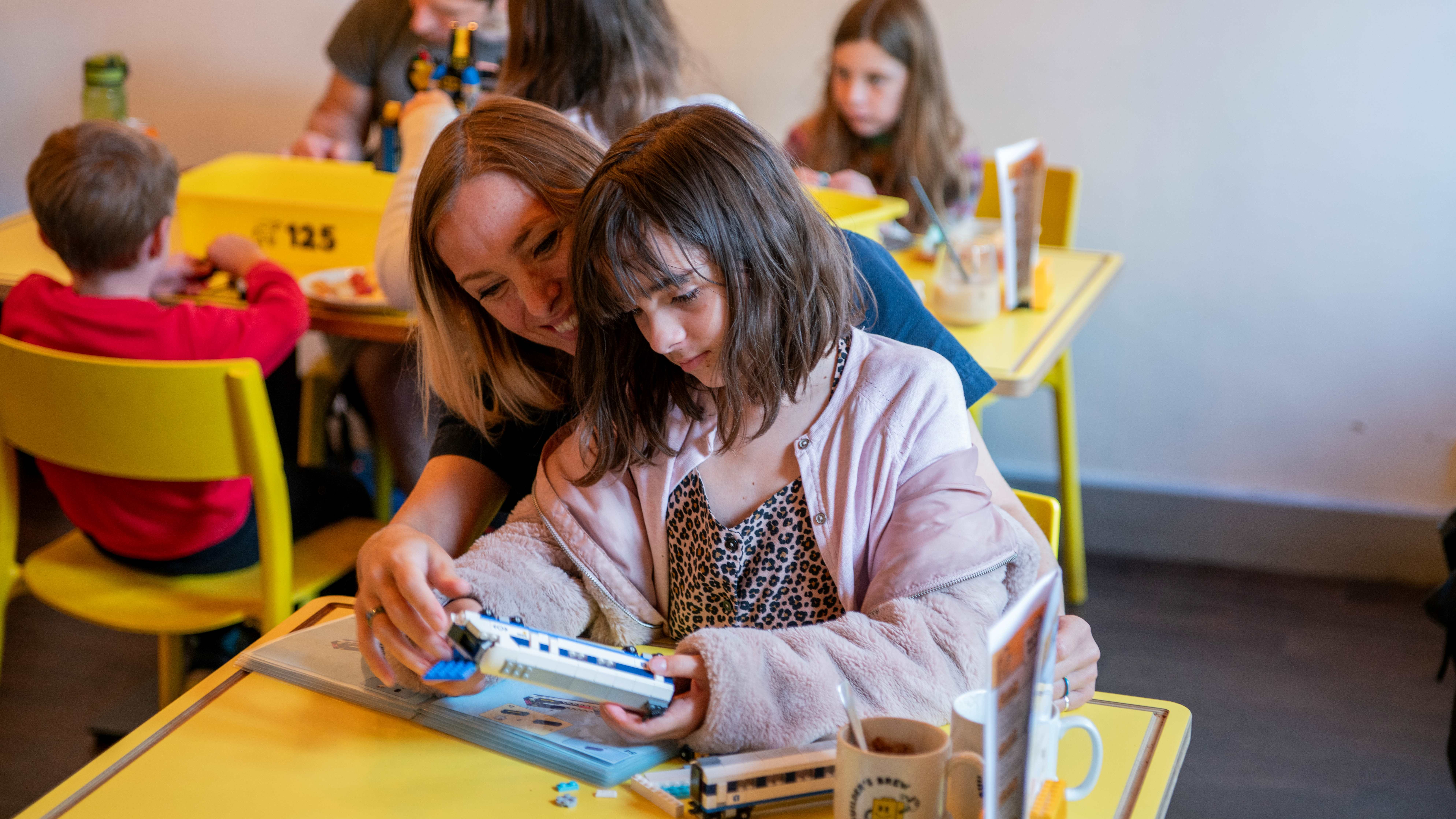 Two people sit at a bright yellow table in a café, assembling a toy train model. Other tables with children and adults are visible in the background, along with mugs and menus on the table, creating a playful and family-friendly atmosphere.