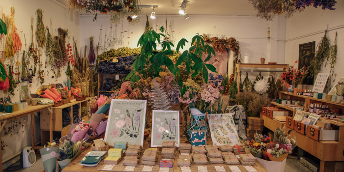 Interior of a boutique shop filled with dried flowers, plants, and botanical arrangements. A central wooden table displays illustrated floral prints, seed packets, and small items, while shelves and walls around the room are decorated with hanging bunches of dried flowers and greenery. The space has warm lighting and a natural, rustic aesthetic.